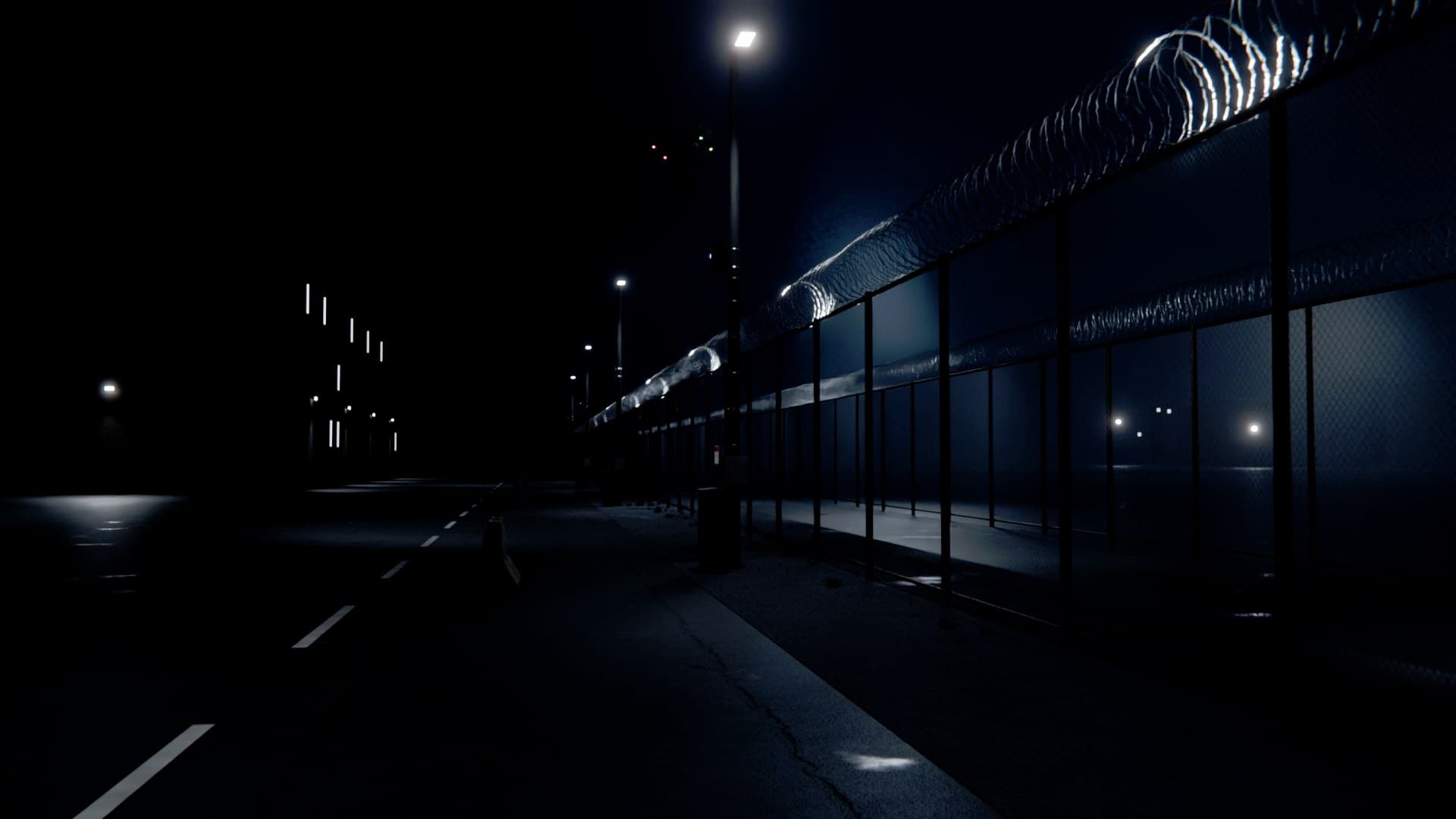 A prison's perimeter road at night, a lit double fence with razor wire, a small drone above it